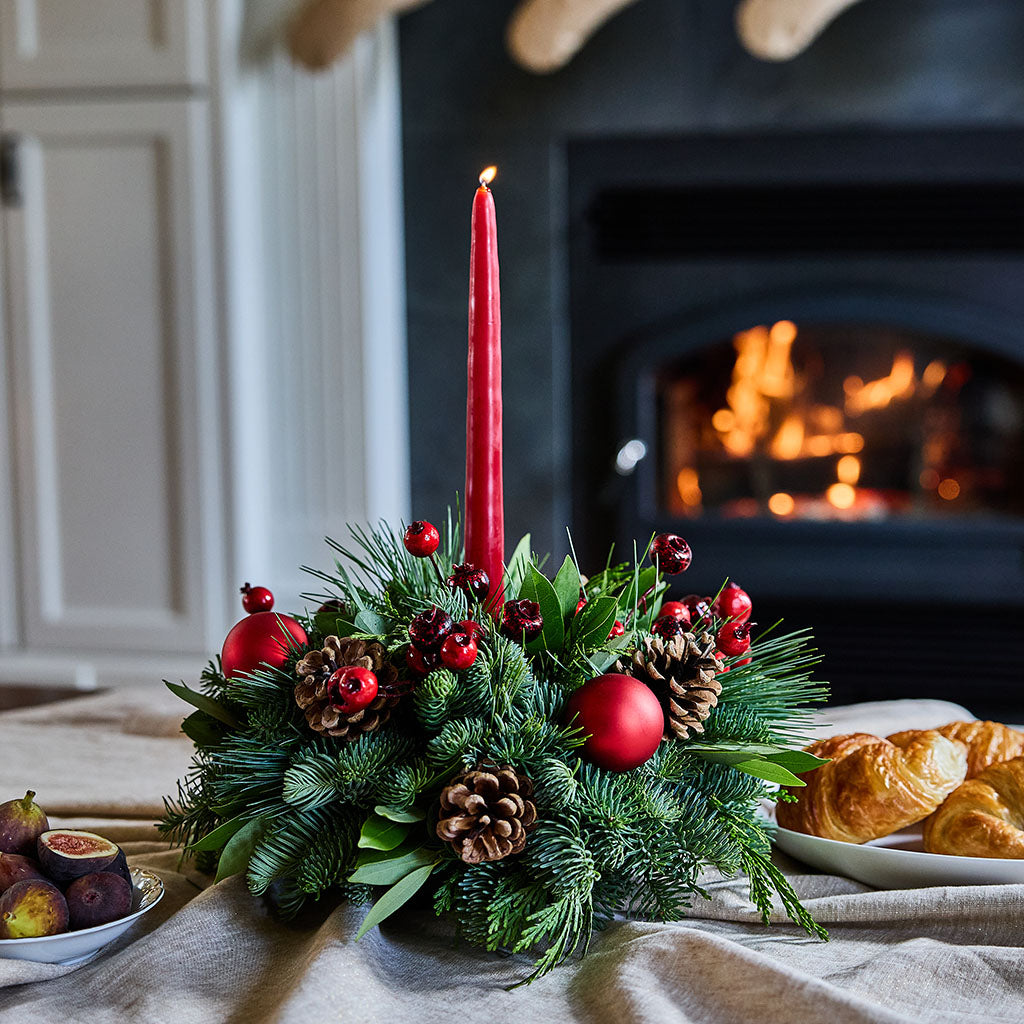 A festive Christmas centerpiece with a tall red taper candle, green pine branches, pinecones, and red ornaments, styled on a table in front of a burning fireplace with croissants and figs nearby.
