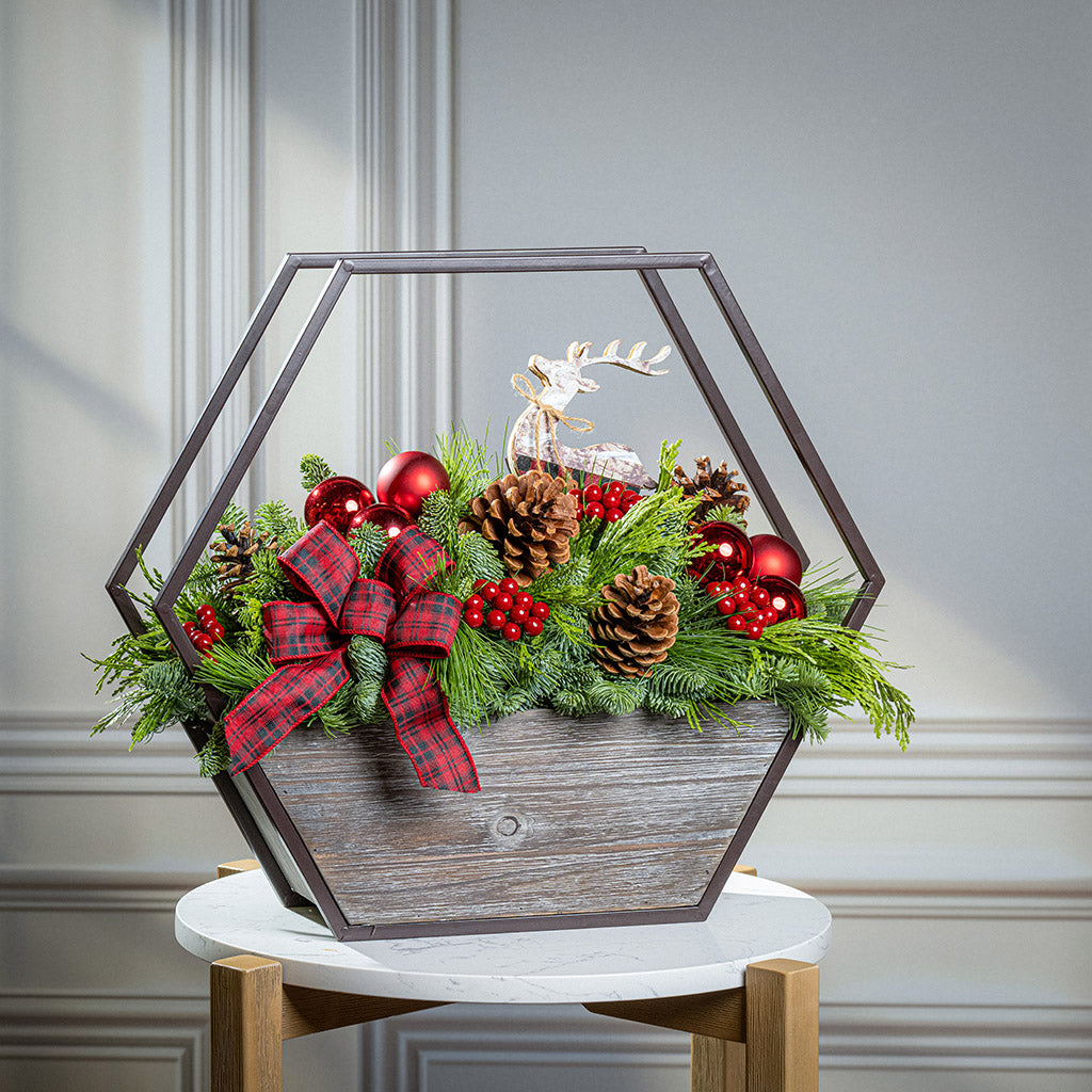 Decorative Christmas arrangement with greenery, red ornaments, and a plaid bow in a geometric wooden box.