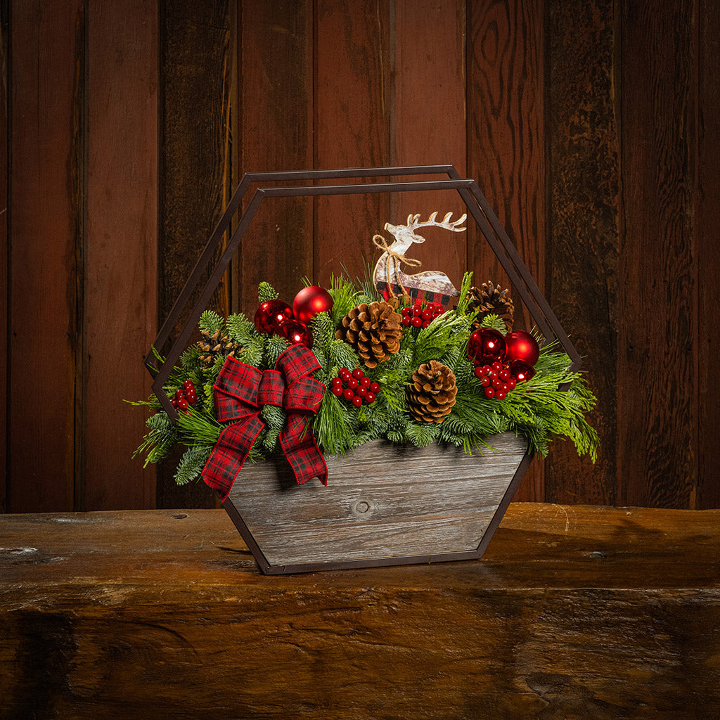 Decorative Christmas arrangement with pine cones, red berries, and a plaid bow in a wooden basket on a wooden surface.