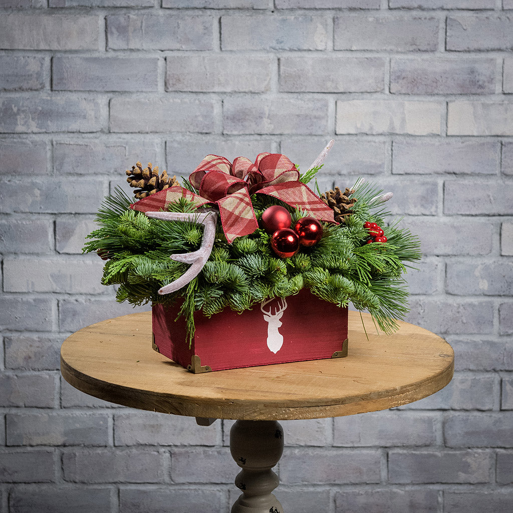 An evergreen arrangement of noble fir, incense cedar, and white pine with red ball clusters, red berry clusters, pinecones, faux antlers, and a linen burgundy plaid bow in a rustic red wooden container with a deer head silhouette