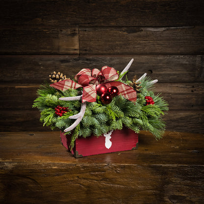 An evergreen arrangement of noble fir, incense cedar, and white pine with red ball clusters, red berry clusters, pinecones, faux antlers, and a linen burgundy plaid bow in a rustic red wooden container with a deer head silhouette