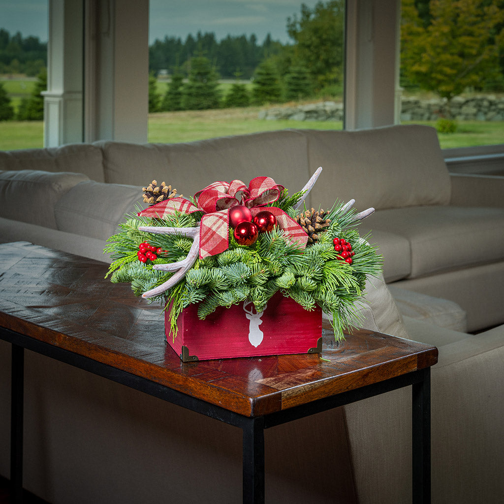 An evergreen arrangement of noble fir, incense cedar, and white pine with red ball clusters, red berry clusters, pinecones, faux antlers, and a linen burgundy plaid bow in a rustic red wooden container with a deer head silhouette