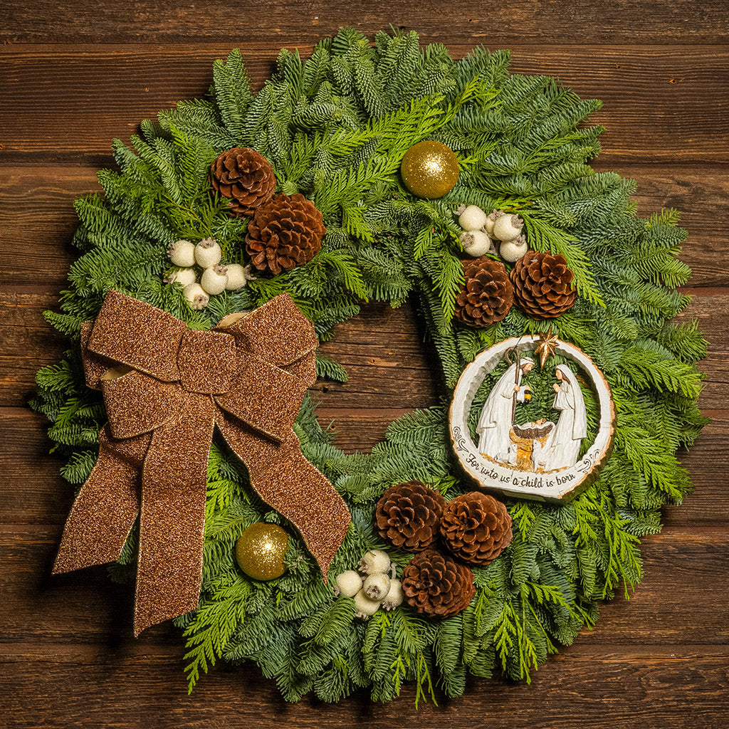 Decorative Christmas wreath with a brown bow, pinecones, and a nativity scene ornament on a wooden background.