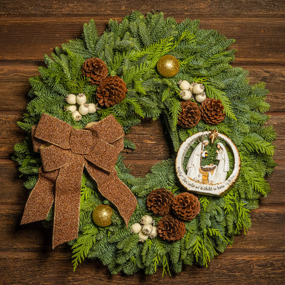 Decorative Christmas wreath with a brown bow, pinecones, and a nativity scene ornament on a wooden background.