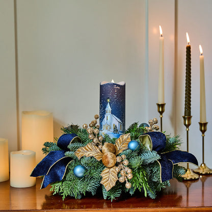 A festive Christmas centerpiece on a wooden table featuring a tall cylindrical candle with a snowy church illustration, surrounded by green pine branches, navy blue ribbons, gold decorative leaves, blue ornaments, and small berries. Lit candles in holders are arranged in the background against a soft beige wall.