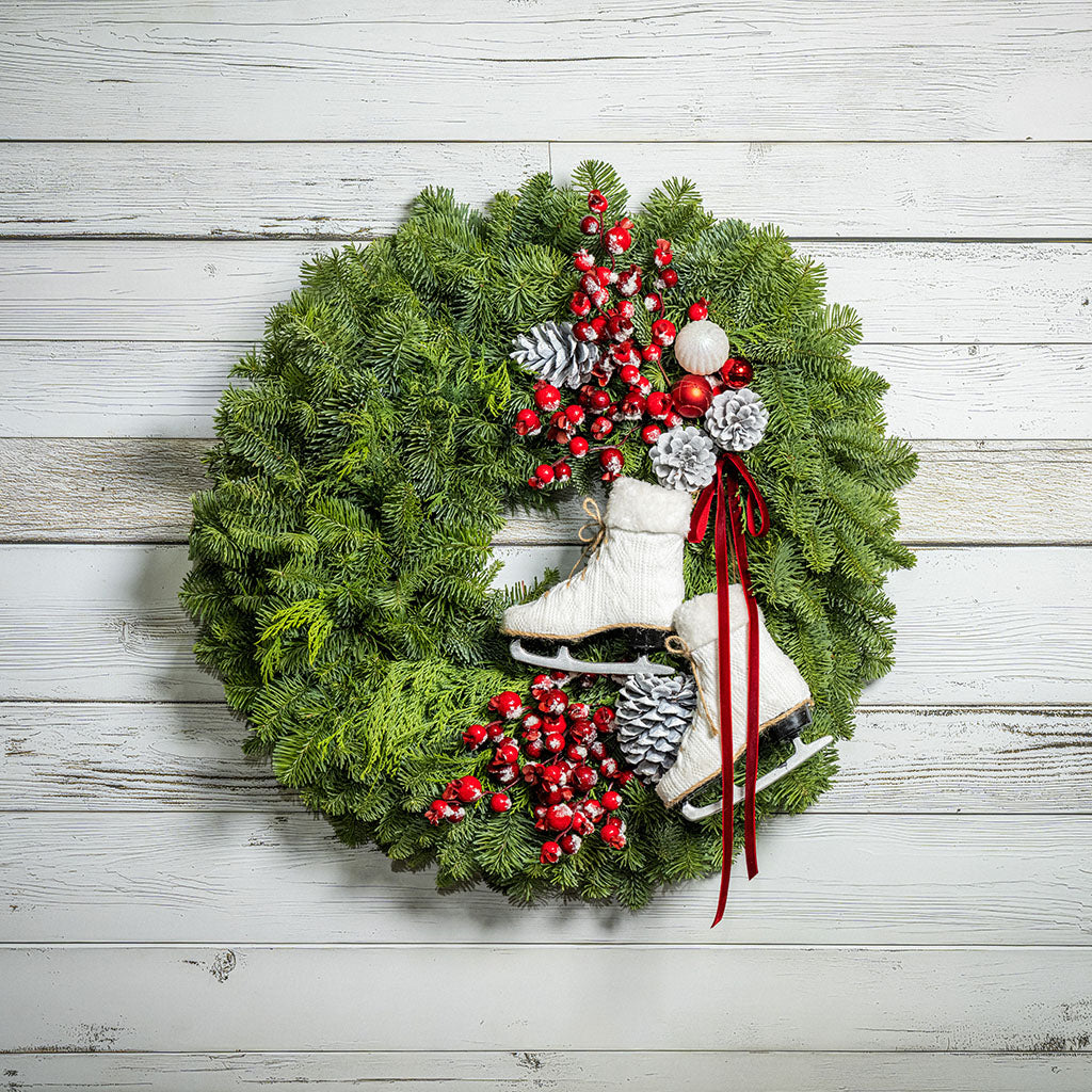 A festive Christmas wreath made of green pine branches, decorated with red berries, white and silver ornaments, pinecones, and two white ice skates, hanging against a white wood plank wall.