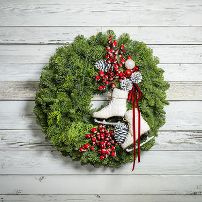 A festive Christmas wreath made of green pine branches, decorated with red berries, white and silver ornaments, pinecones, and two white ice skates, hanging against a white wood plank wall.