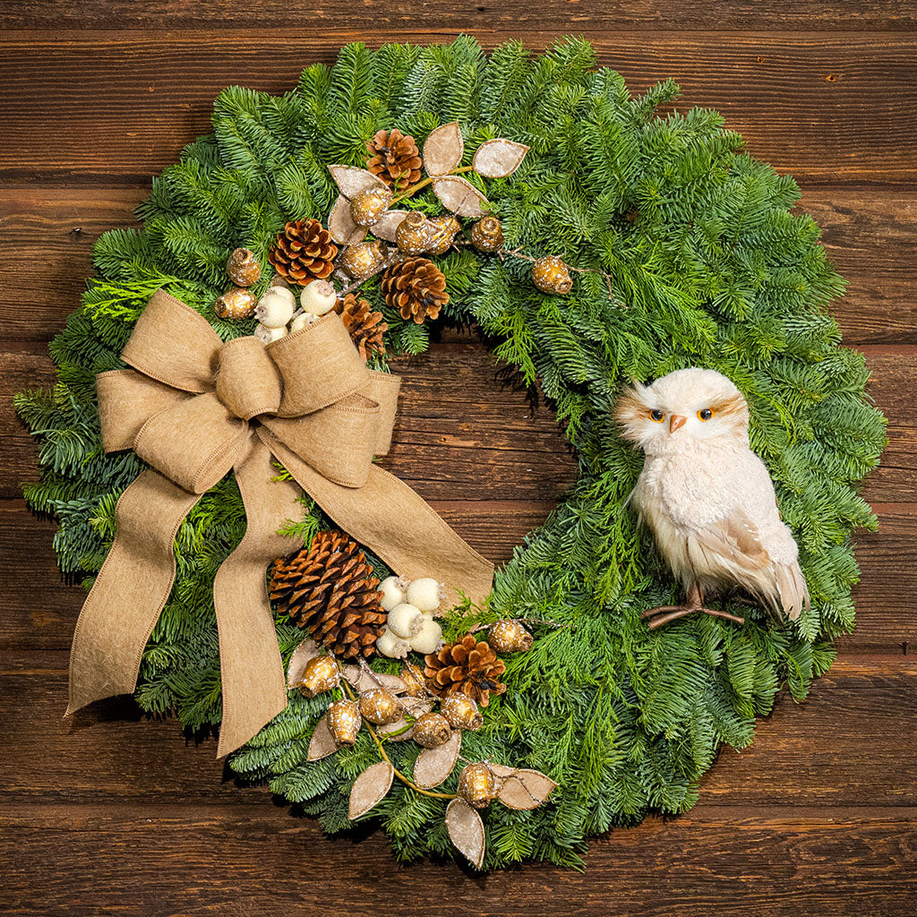 A festive Christmas wreath made of green pine branches, decorated with a large tan burlap bow, pinecones, gold berries, white berries, and a small white owl ornament, displayed on a dark wood background.