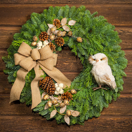 A festive Christmas wreath made of green pine branches, decorated with a large tan burlap bow, pinecones, gold berries, white berries, and a small white owl ornament, displayed on a dark wood background.