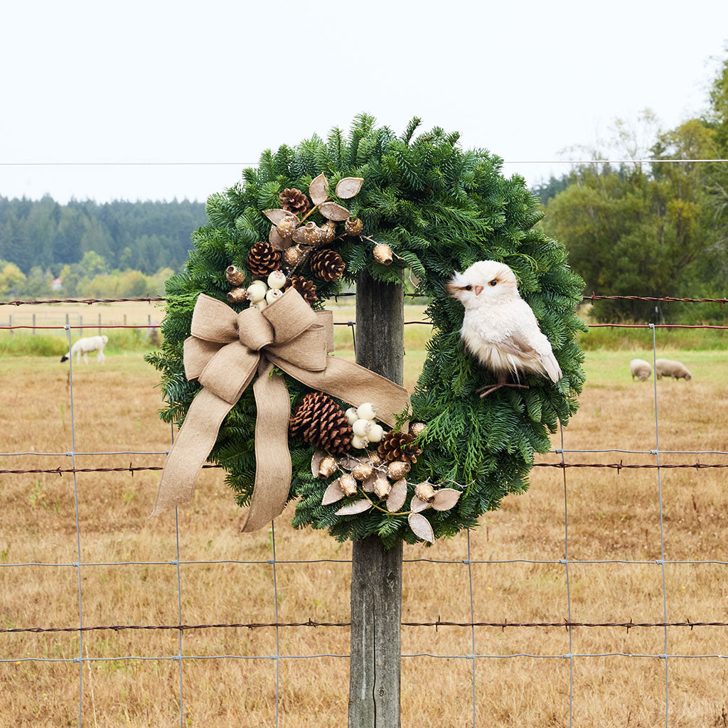 A rustic green pine wreath with a large tan burlap bow, brown pinecones, dried flowers, and a white bird ornament, hanging on a wooden fence post in an outdoor field.