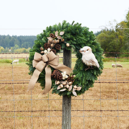 A rustic green pine wreath with a large tan burlap bow, brown pinecones, dried flowers, and a white bird ornament, hanging on a wooden fence post in an outdoor field.