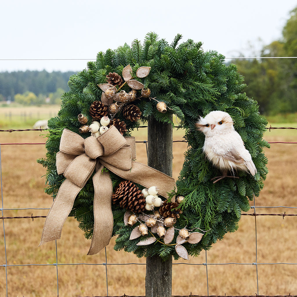 A green pine wreath hanging on a wire fence outdoors, decorated with a large tan burlap bow, pinecones, dried leaves, and white berries, with a small white bird perched on the right side.