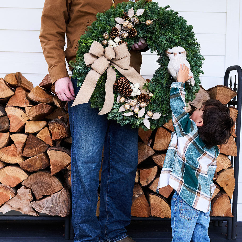 A large green pine wreath with a beige burlap bow, pinecones, and white berries is held by an adult standing in front of a stacked wood fire. A child in a teal plaid shirt reaches up to touch a white owl ornament attached to the wreath.