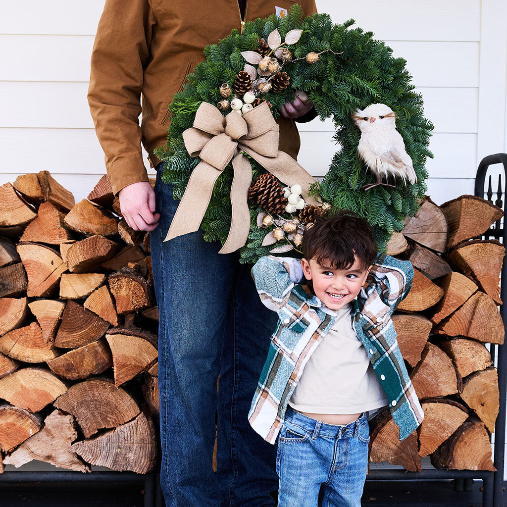 A festive evergreen wreath with a large beige ribbon bow, pinecones, berries, and a white bird ornament, held by an adult and a smiling child in front of stacked firewood.