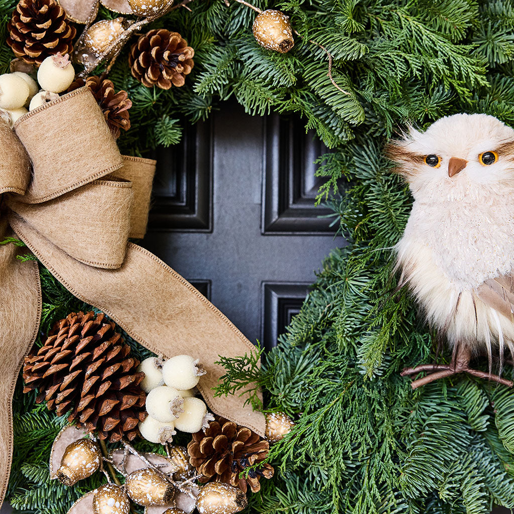 A festive Christmas wreath made of green pine branches, decorated with a large tan burlap bow, pinecones, small white berries, gold-beaded ornaments, and a fluffy cream owl decoration, arranged in front of a dark paneled door.