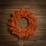 A deep orange wreath made of preserved orange myrtle, preserved eucalyptus, and orange flax pods hanging on a wood background. 