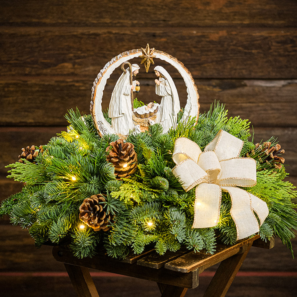 An evergreen arrangement of noble fir, cedar, and pine with natural pinecones, ivory bows, and a nativity scene decoration sitting on a wooden table with a dark wood background.