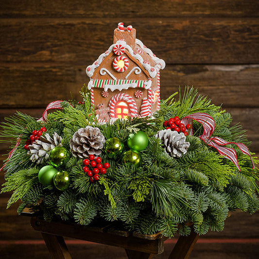 An evergreen arrangement of noble fir, cedar, and pine with green ball ornaments, faux red berries, frosted pinecones, two striped bow tucks, and a gingerbread house decoration with a dark wood background