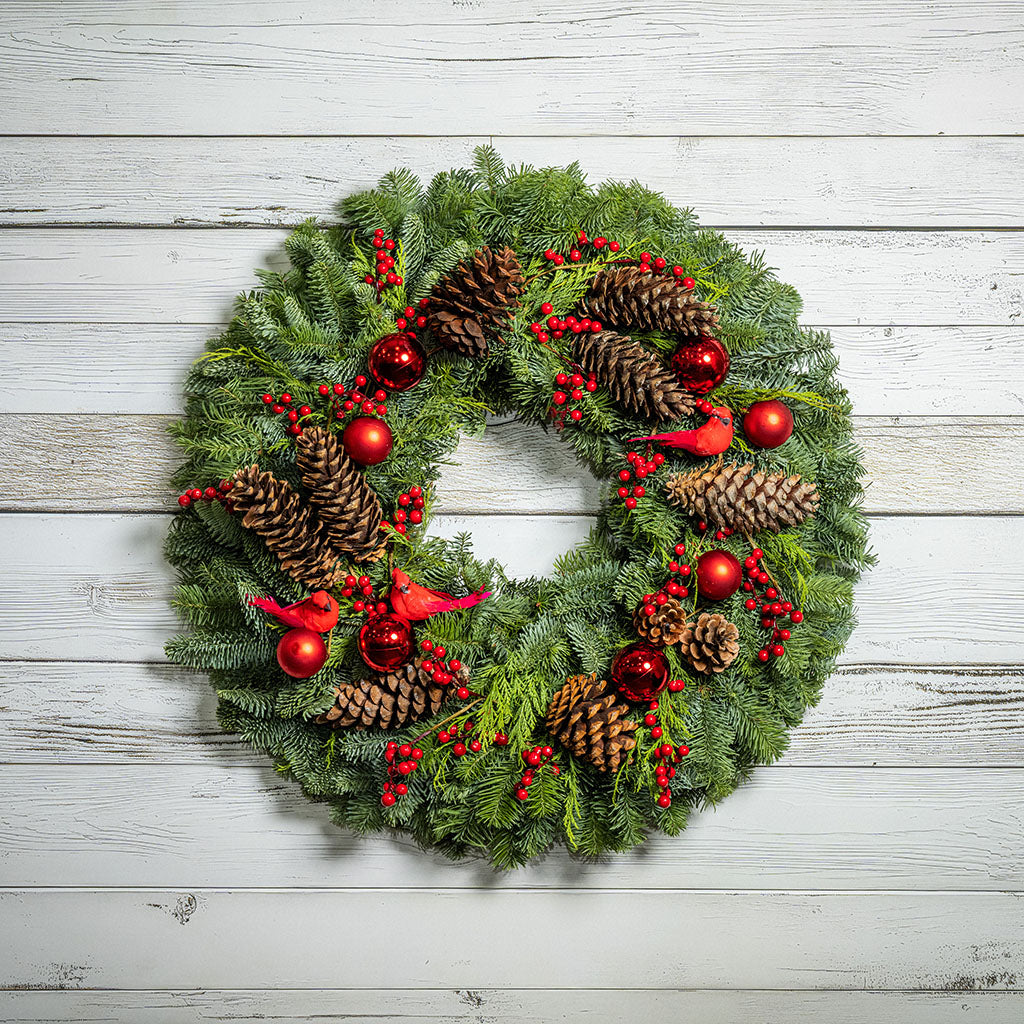 A festive Christmas wreath made of green pine branches, decorated with brown pinecones, red berries, red ornaments, and small red birds, displayed against a white wood plank background.