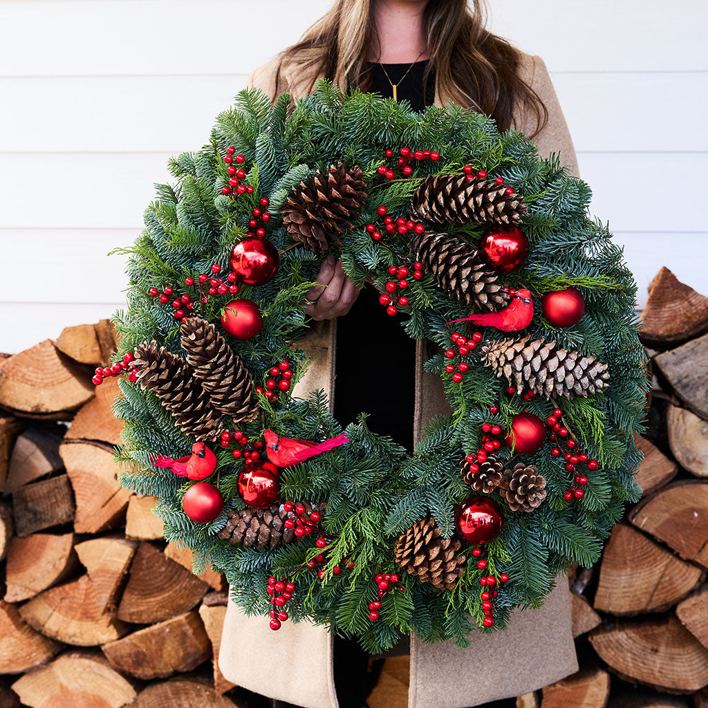A person holds a large green Christmas wreath decorated with pinecones, red berries, shiny red ornaments, and small red bird ornaments, positioned in front of a stack of firewood and a white paneled wall.