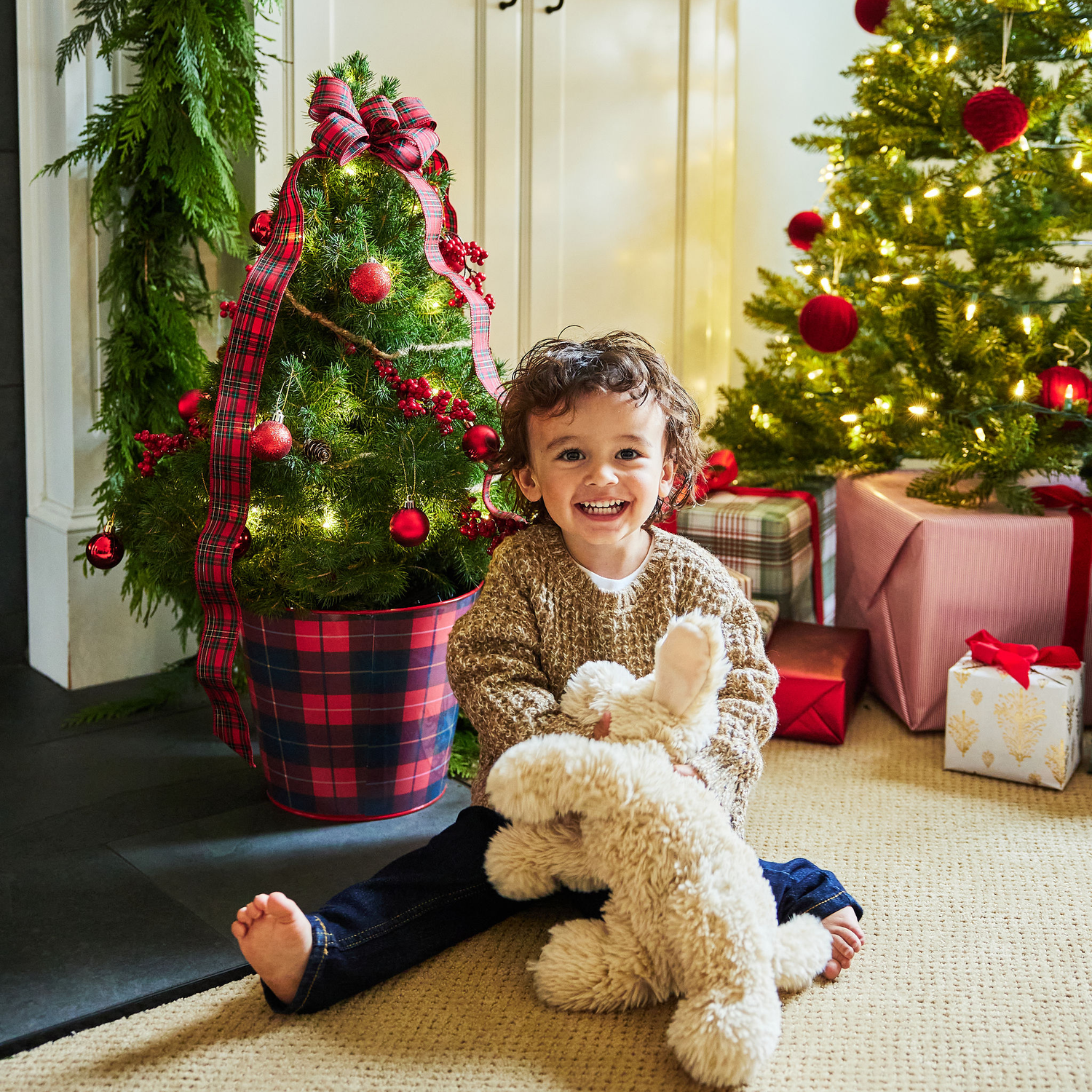 Child sitting on the floor with a teddy bear in front of a decorated Christmas tree.