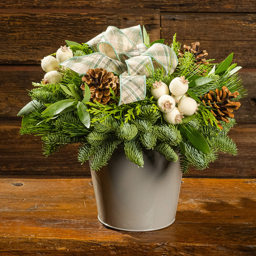 Decorative floral arrangement with greenery, white berries, and pine cones in a gray pot on a wooden surface.