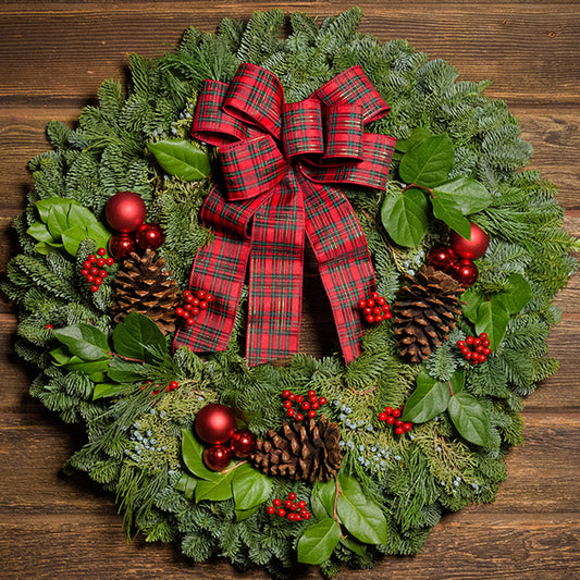 An arrangement of noble fir, incense cedar, juniper, and salal leaves with ponderosa pinecones, faux red berry clusters, red ball ornaments, and a plaid bow on a dark wood background.