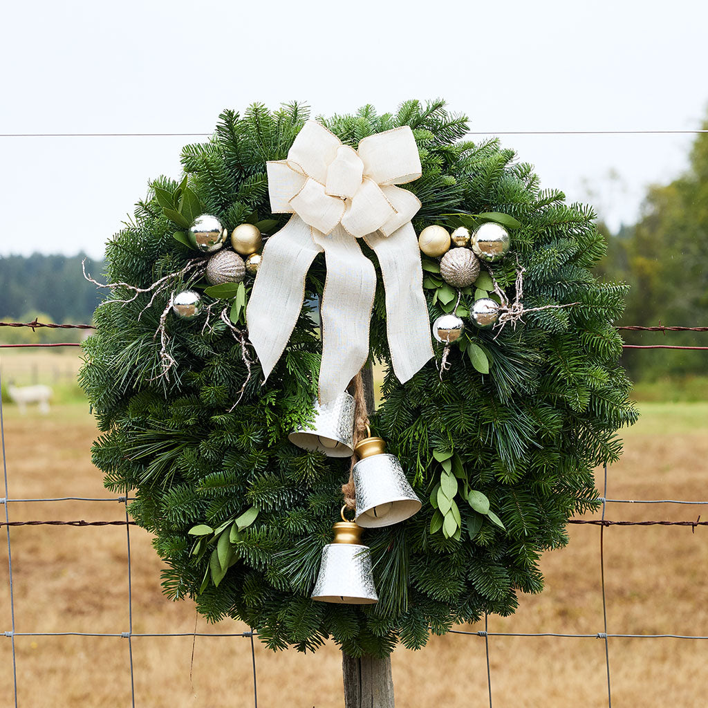 A green pine wreath decorated with a large cream ribbon bow, silver bell ornaments, and small silver and gold baubles, hanging on a wire fence in a rural outdoor setting.