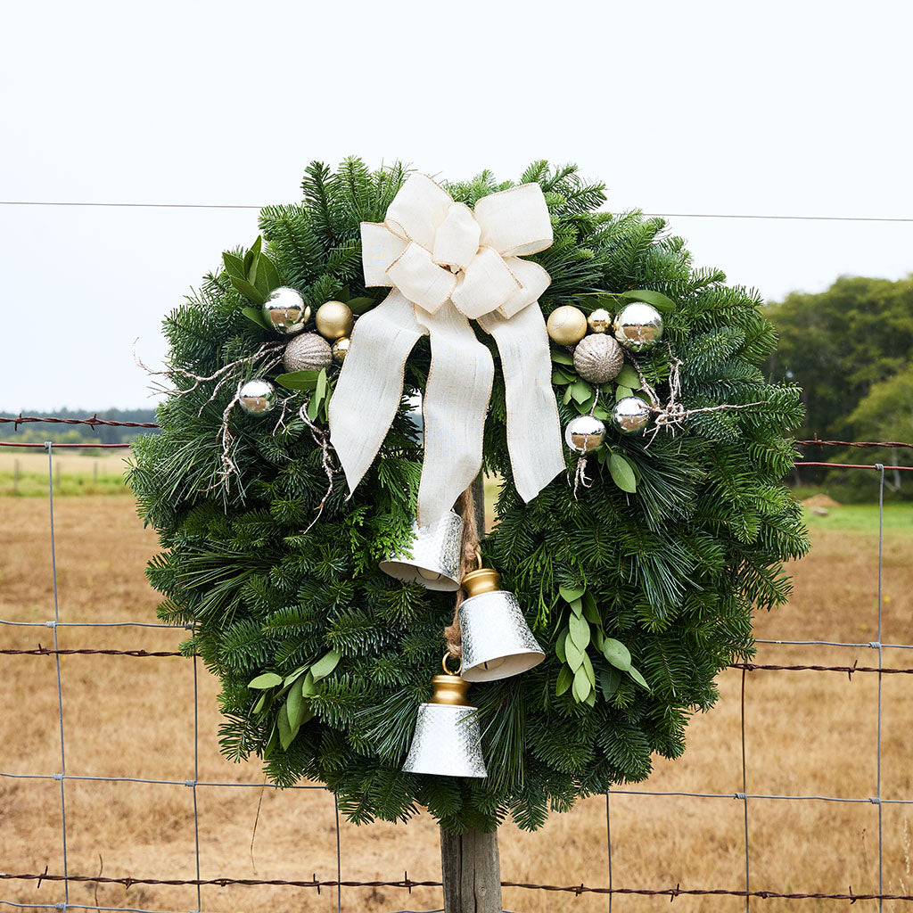 A green evergreen wreath hanging on a wire fence, decorated with a large cream bow, silver and gold ornaments, and two silver bells, with a dry field and trees in the background.