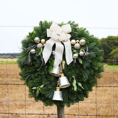 A green evergreen wreath hanging on a wire fence, decorated with a large cream bow, silver and gold ornaments, and two silver bells, with a dry field and trees in the background.
