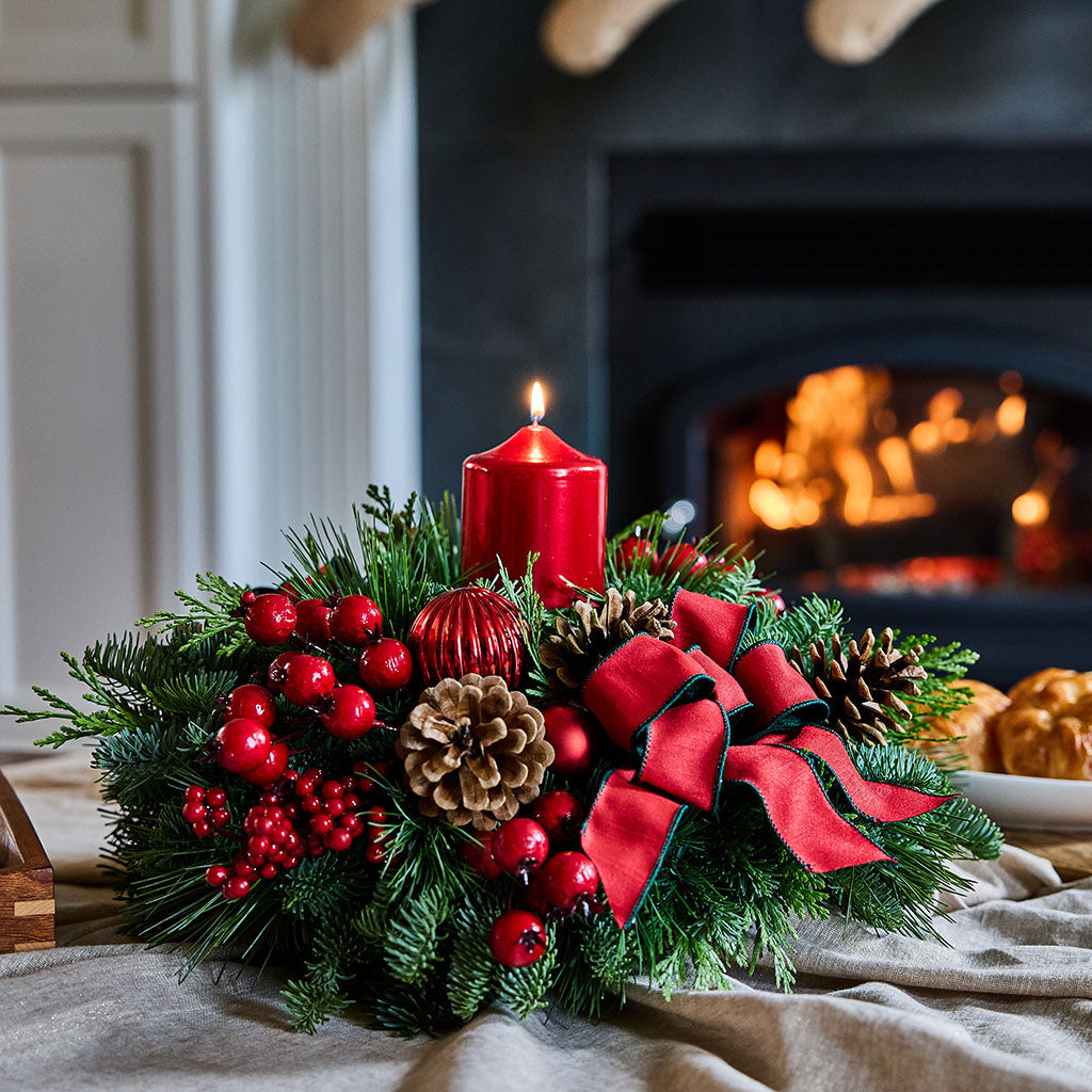 A festive Christmas wreath centerpiece with green pine branches, red berries, pinecones, a large red bow, and a lit red pillar candle, styled on a table in front of a warm fireplace.
