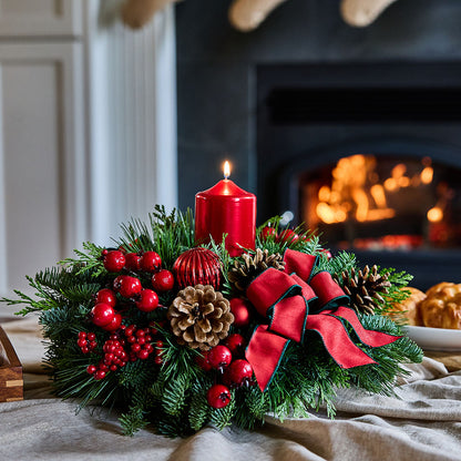 A festive Christmas wreath centerpiece with green pine branches, red berries, pinecones, a large red bow, and a lit red pillar candle, styled on a table in front of a warm fireplace.