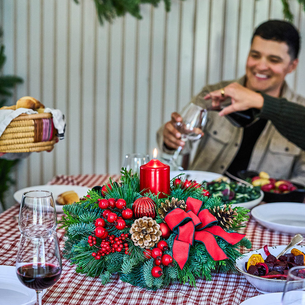 A festive Christmas table centerpiece with green pine branches, red berries, pinecones, and a red bow, set on a red-and-white checkered tablecloth. A man in a beige jacket is blurred in the background pouring wine from a bottle into a glass.