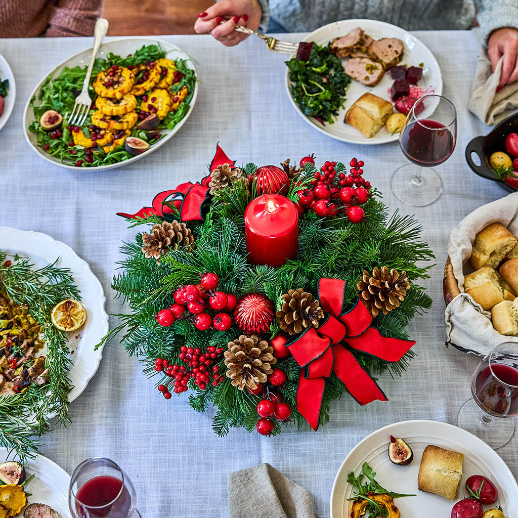 A festive Christmas centerpiece on a dining table, featuring a green pine wreath with red ribbons and bows, red berries, pinecones, and a central red candle. Surrounding plates of food, wine glasses, and hands at the table are visible, indicating a lifestyle setting.