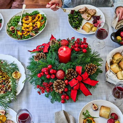 A festive Christmas centerpiece on a dining table, featuring a green pine wreath with red ribbons and bows, red berries, pinecones, and a central red candle. Surrounding plates of food, wine glasses, and hands at the table are visible, indicating a lifestyle setting.