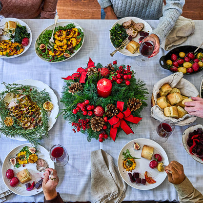Overhead view of a festive holiday dinner table with multiple white plates of food, red wine glasses, and a central Christmas centerpiece made of greenery, red bows, pinecones, and a lit red candle. Hands holding forks and wine glasses are visible around the table, and the scene is set in a real dining environment.