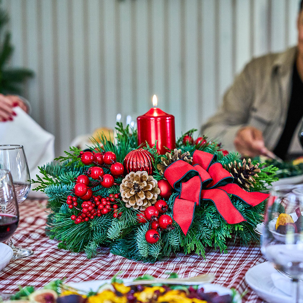 A festive Christmas table centerpiece with green pine branches, red berries, a lit red pillar candle, pinecones, and a large red bow, set on a red-and-white checkered tablecloth with blurred people and dining glasses in the background.