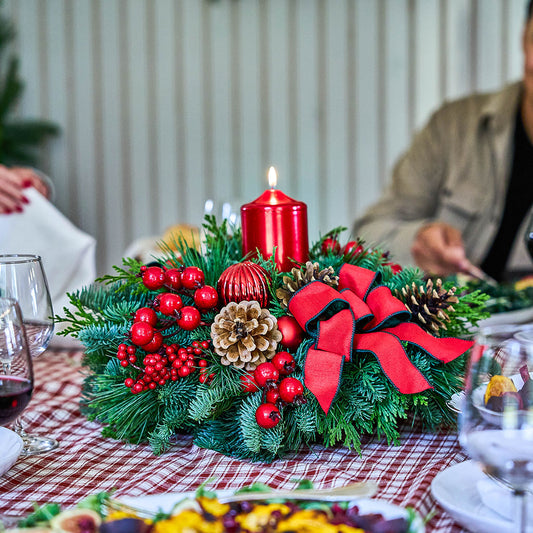 A festive Christmas table centerpiece with green pine branches, red berries, a lit red pillar candle, pinecones, and a large red bow, set on a red-and-white checkered tablecloth with blurred people and dining glasses in the background.