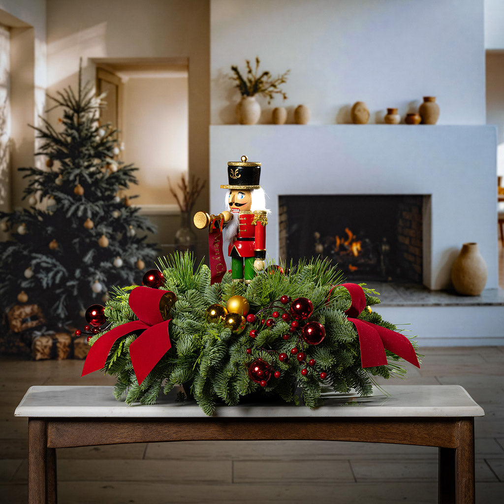 table centerpiece with greenery with red and gold ball ornaments, faux red berry branches, red velvet bows, and a wooden bugler Nutcracker decoration on a table in front of Christmas tree and fireplace.