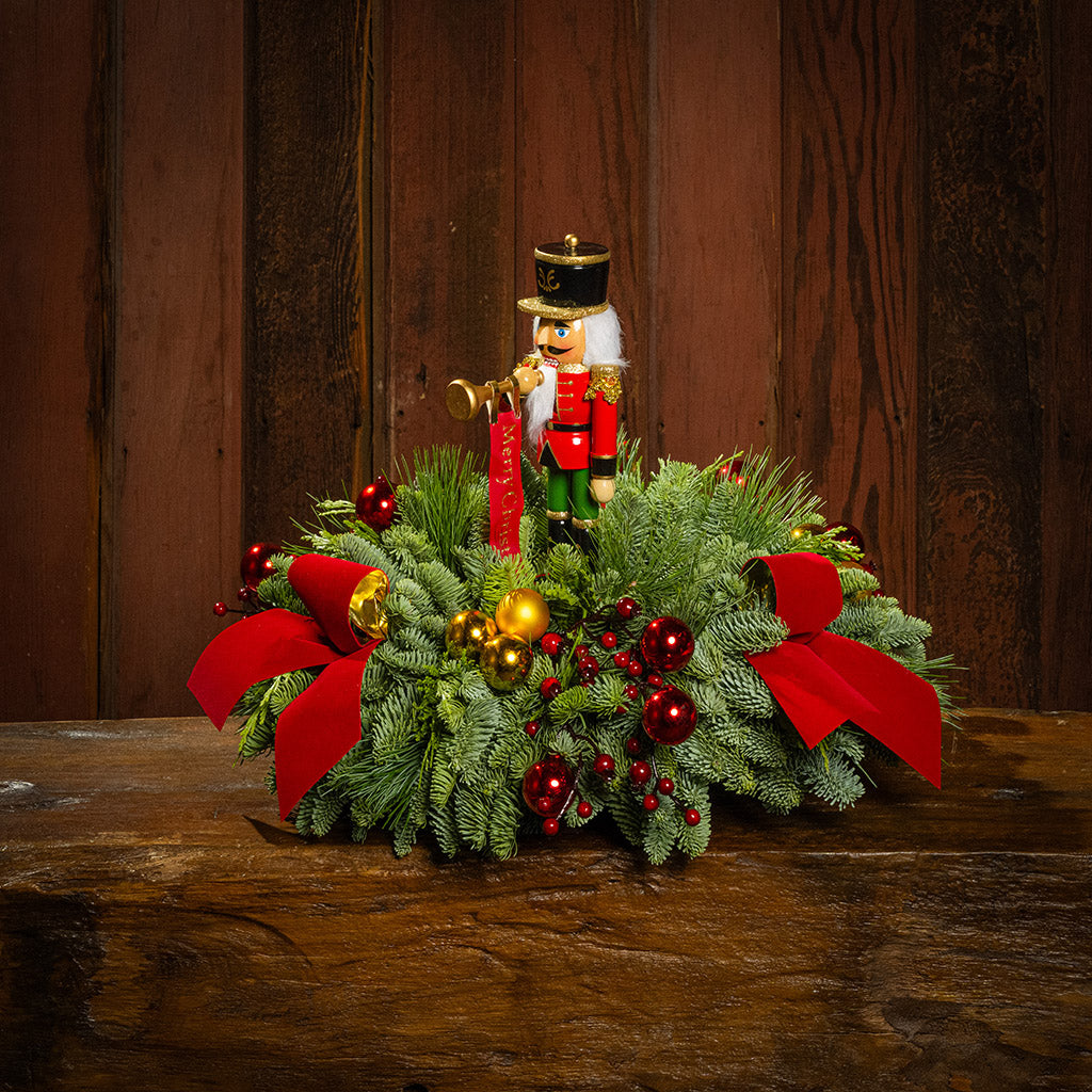 greenery with red and gold ball ornaments, faux red berry branches, red velvet bows, and a wooden bugler Nutcracker decoration on a wooden table with a wood background.