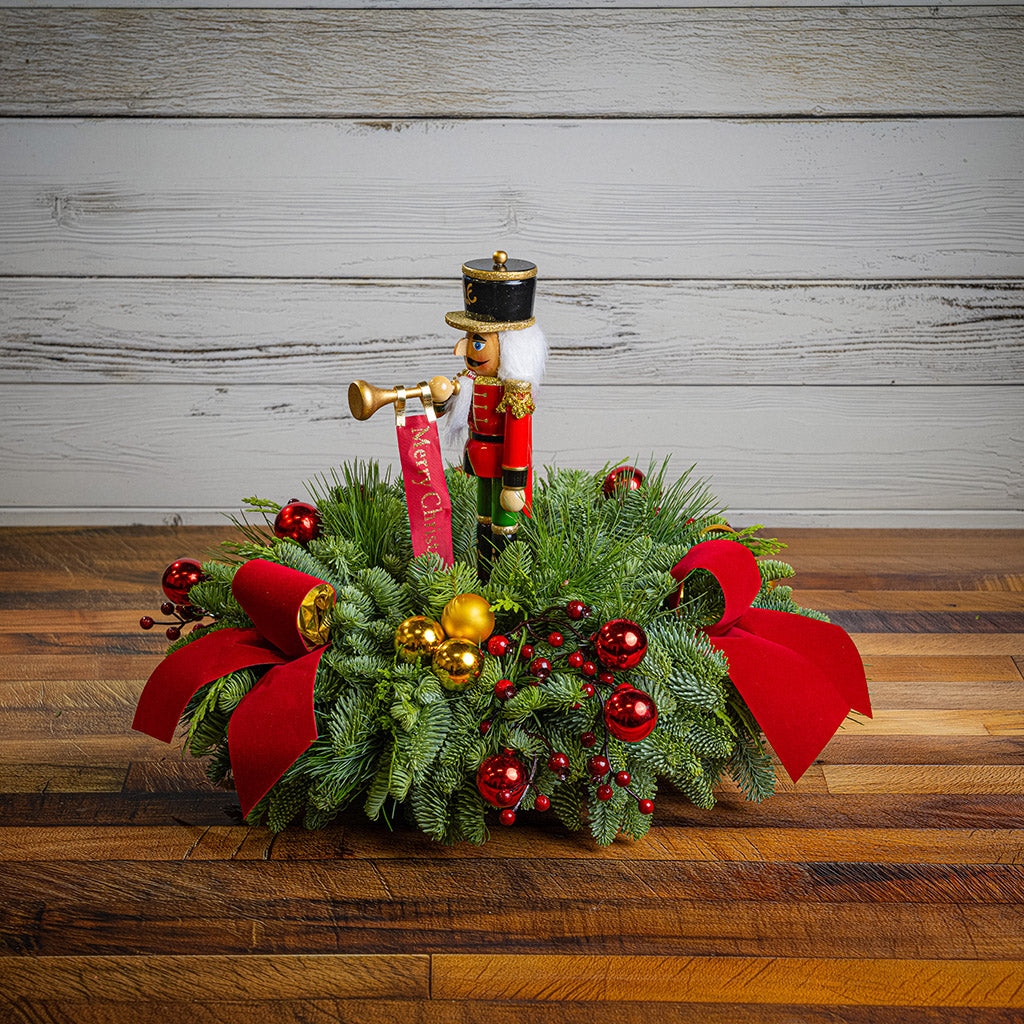 greenery with red and gold ball ornaments, faux red berry branches, red velvet bows, and a wooden bugler Nutcracker decoration on a wood table with a white wooden background.