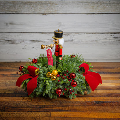 greenery with red and gold ball ornaments, faux red berry branches, red velvet bows, and a wooden bugler Nutcracker decoration on a wood table with a white wooden background.