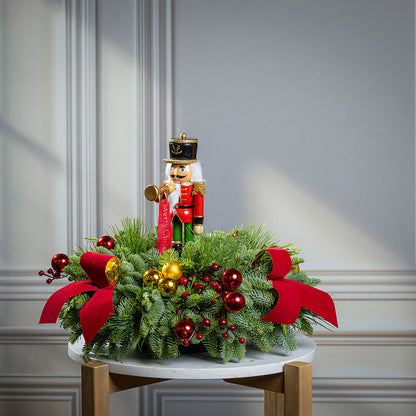 greenery with red and gold ball ornaments, faux red berry branches, red velvet bows, and a wooden bugler Nutcracker decoration on a marble table.