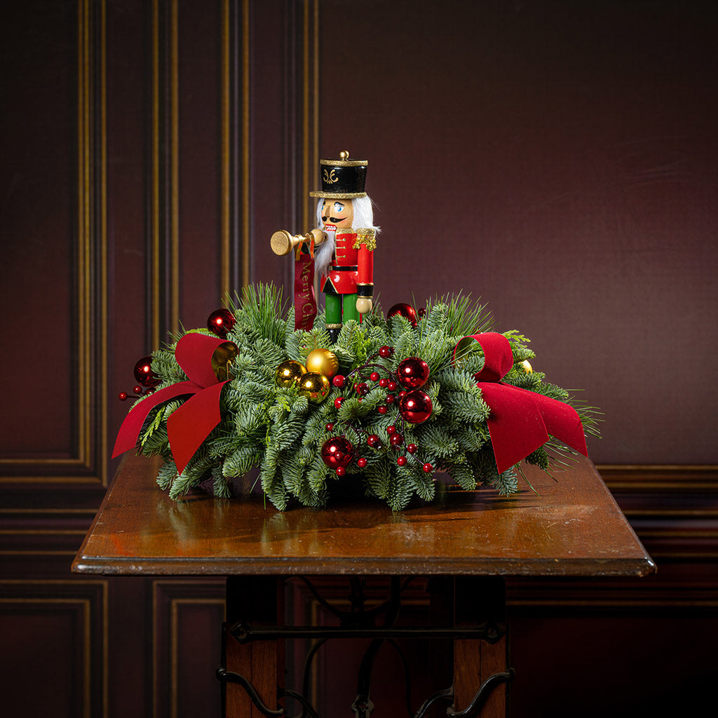 greenery with red and gold ball ornaments, faux red berry branches, red velvet bows, and a wooden bugler Nutcracker decoration on a wooden table.