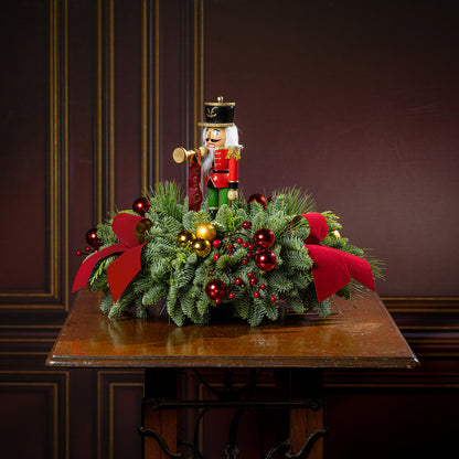 greenery with red and gold ball ornaments, faux red berry branches, red velvet bows, and a wooden bugler Nutcracker decoration on a wooden table.