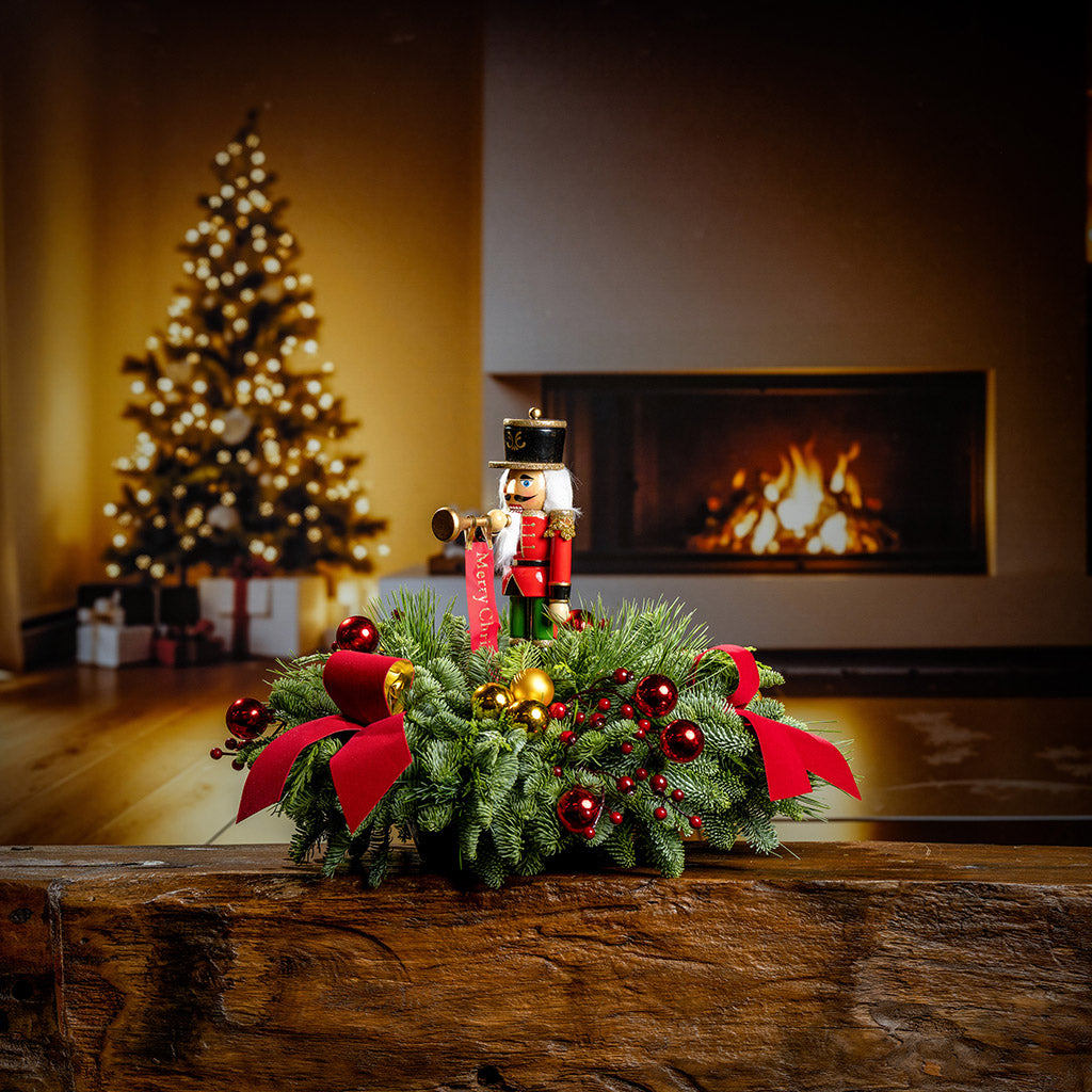 greenery with red and gold ball ornaments, faux red berry branches, red velvet bows, and a wooden bugler Nutcracker decoration on a wooden table in front of fireplace and Christmas tree.