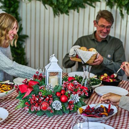 A festive Christmas table centerpiece with red berries, green pine branches, white pinecones, and a white lantern, set on a red-and-white checkered tablecloth. A family is gathered around the table, with a man holding a basket of bread in the background.