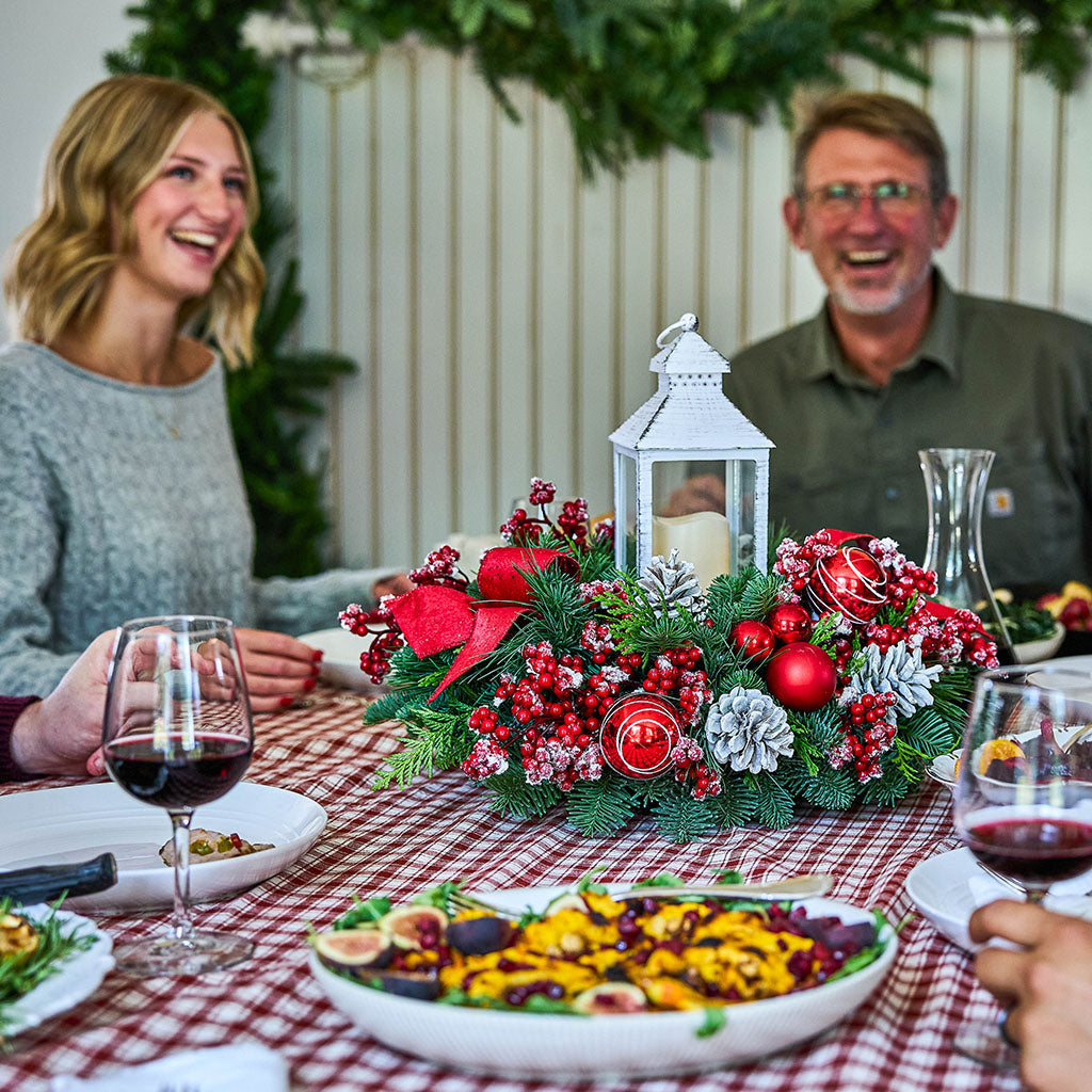 A festive Christmas table centerpiece with red berries, pine branches, silver pinecones, and a white lantern, set on a red-and-white checkered tablecloth with plates of food and wine glasses. A smiling woman is visible on the left, and a man is visible on the right in a real dining environment.