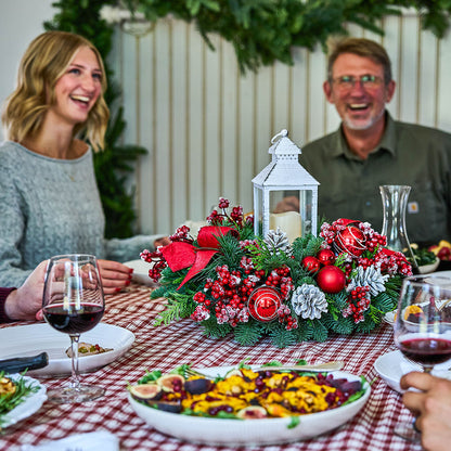 A festive Christmas table centerpiece with red berries, pine branches, silver pinecones, and a white lantern, set on a red-and-white checkered tablecloth with plates of food and wine glasses. A smiling woman is visible on the left, and a man is visible on the right in a real dining environment.