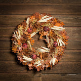 A dried wreath made of cafe colored preserved oak leaves, natural corn husk, amber oak leaves with hints of gold-marbled accents, natural pinecones, and natural ruby pomegranates hung on a dark wood background.